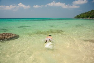 A person swimming in clear turquoise water with a rocky shore and a hill in the background.