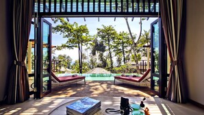 A room with a large window, two red lounge chairs, a table with a book and a camera, and a view of a pool and greenery outside. - Ajinta Resort (Ko Kood)