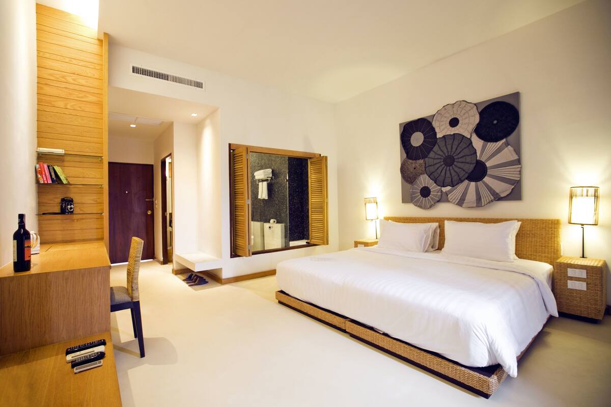 A modern hotel room with a large bed, a wooden desk, a bookshelf, and a wall art piece.