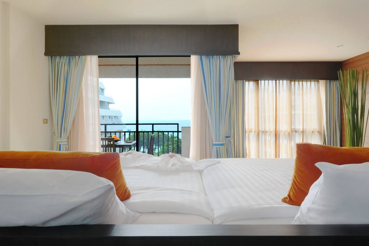 Habitación de hotel con una cama grande, dos almohadas naranjas, vista al mar y un balcón con mesa y sillas.