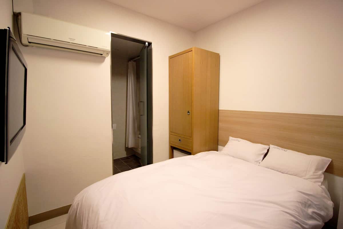 Habitació d'hotel amb un llit, un armari de fusta, una porta que dona a un bany i una unitat de aire condicionat penjada a la paret.