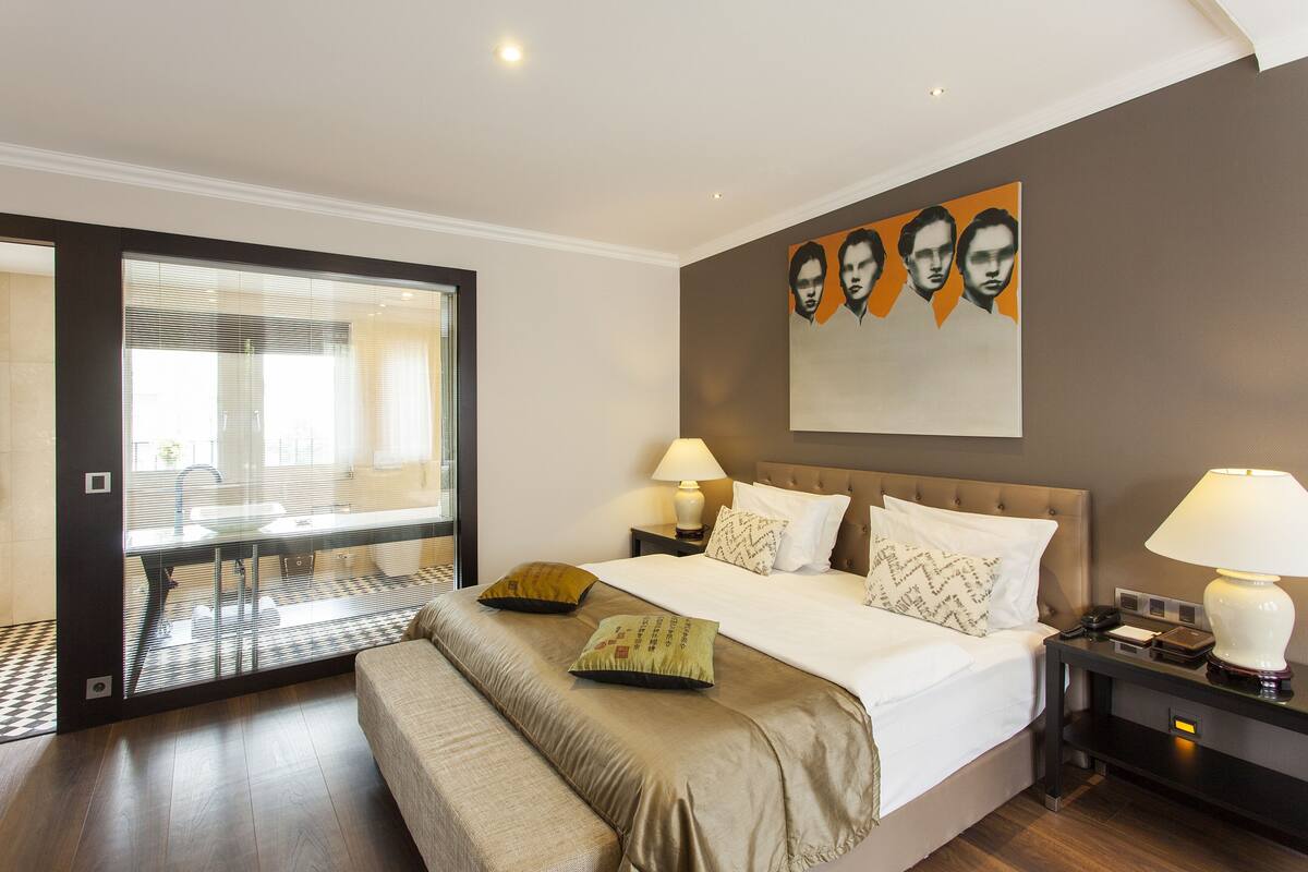 A modern hotel room with a large bed, bedside tables, a glass-enclosed bathroom, and a painting on the wall.
