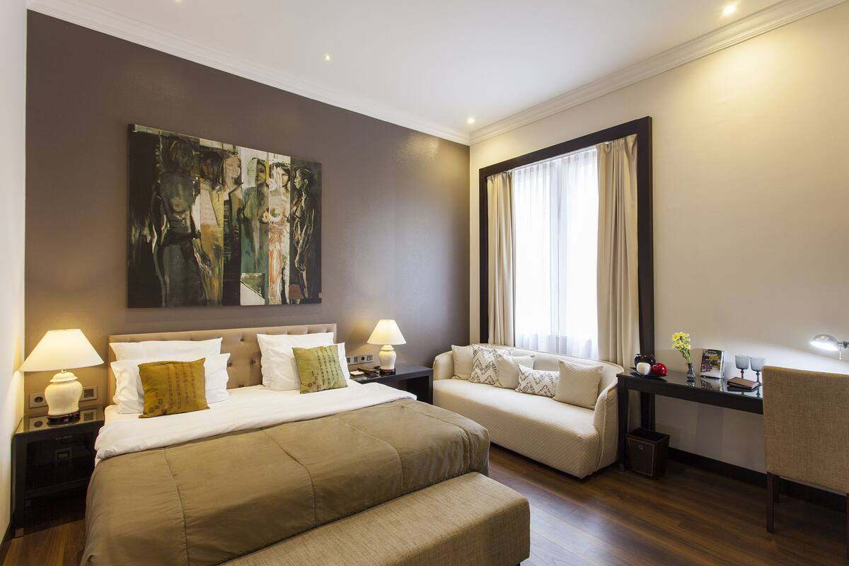 A hotel room with a large bed, a sofa, a desk with a lamp, and a painting on the wall.