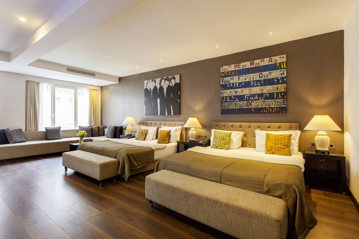 A hotel room with a large bed, two smaller beds, a sofa, and a bench. There are two wall art pieces and a window with curtains.