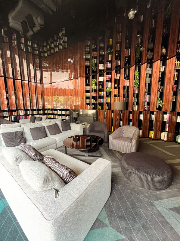 A modern living room with a white sofa, a round coffee table, and a patterned ottoman. There is a bookshelf filled with books and decorative items, and a lamp on a side table.