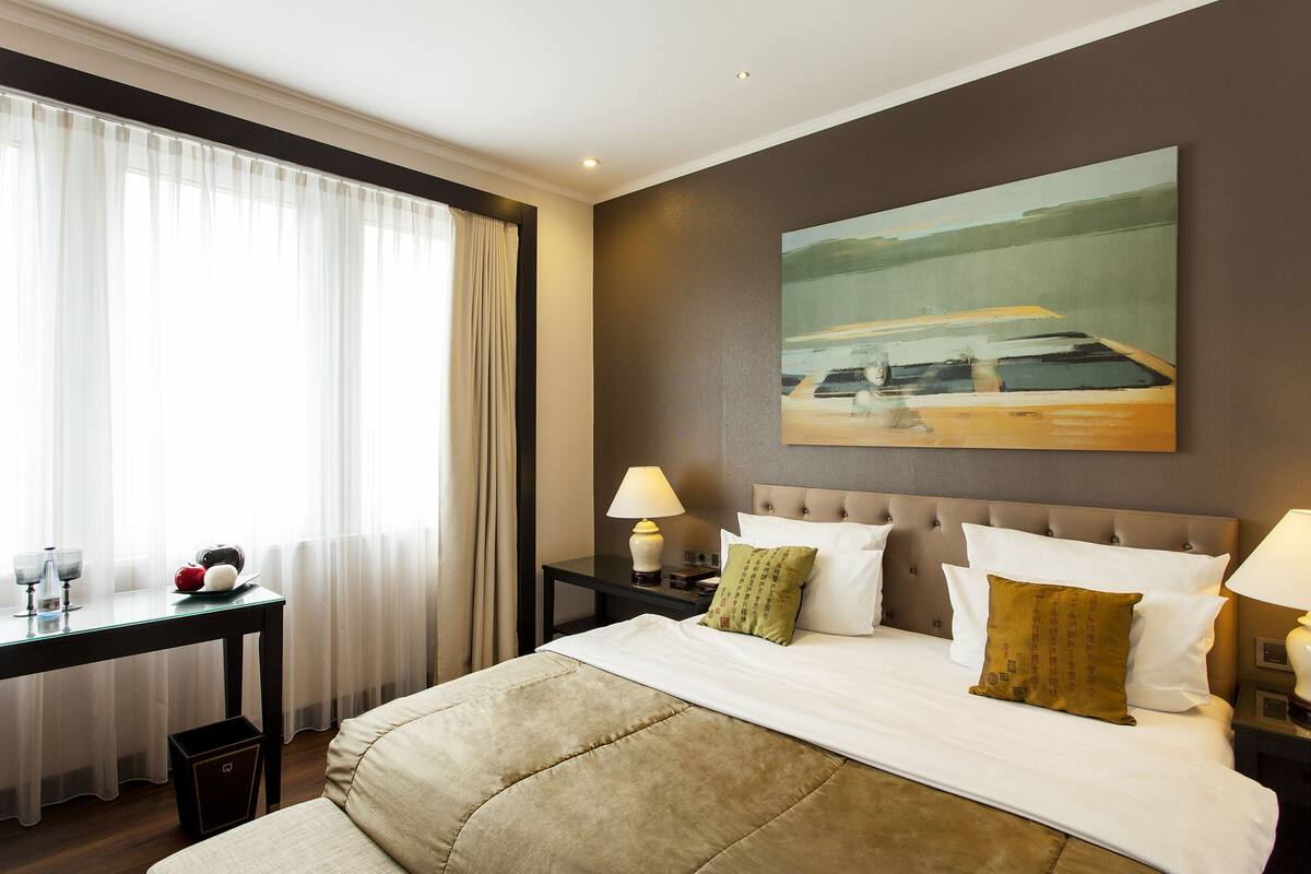 A hotel room with a bed, two bedside tables, a desk with a chair, a window with curtains, and a painting on the wall.