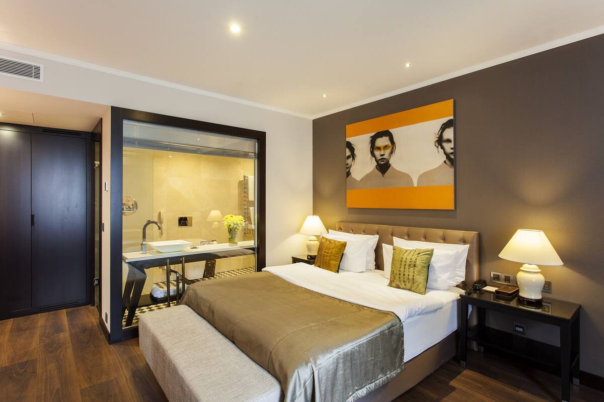 A modern hotel room with a large bed, bedside lamps, a mirror, and a painting on the wall.