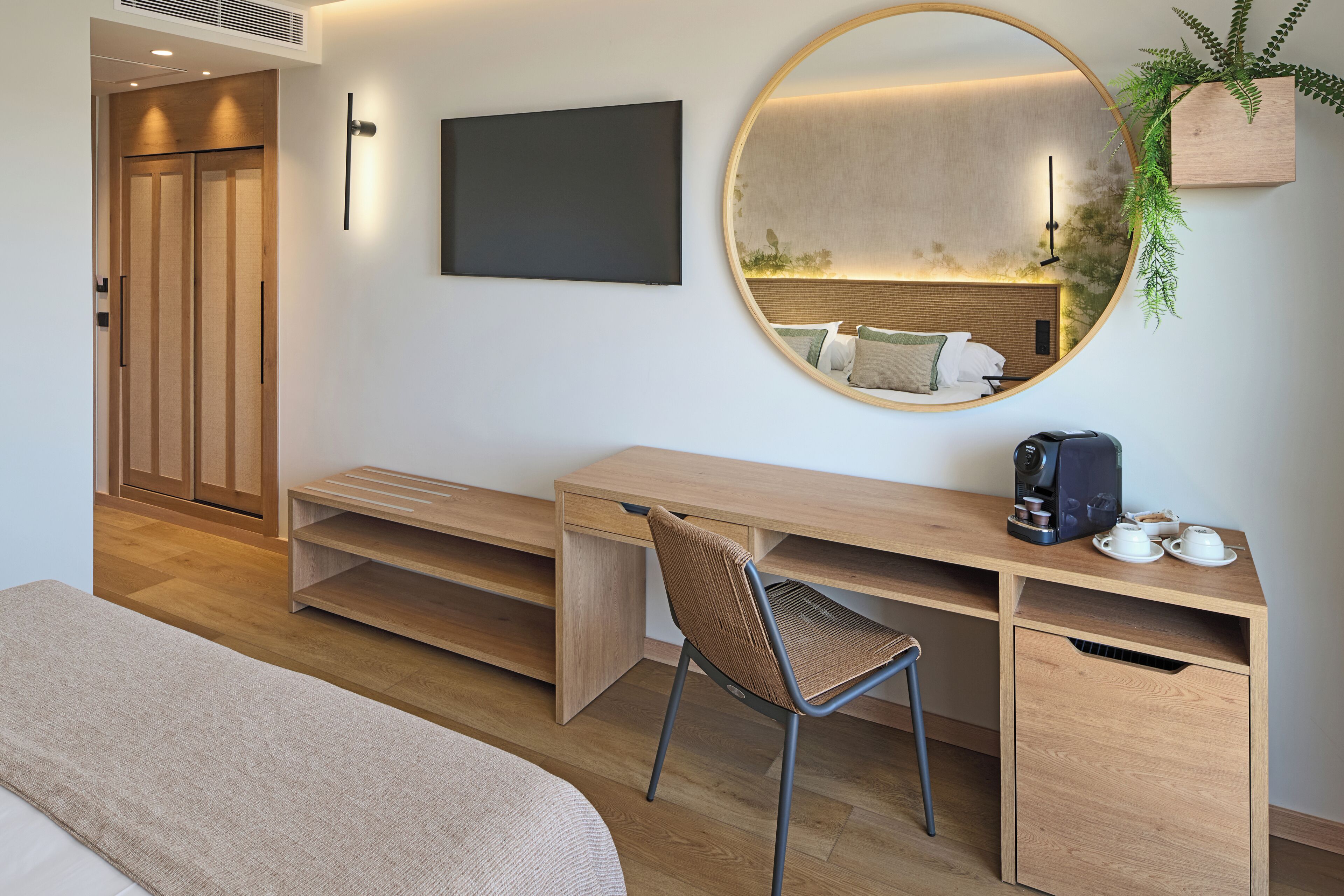 A modern bedroom with a wooden desk, a round mirror, a bed, and a small plant.