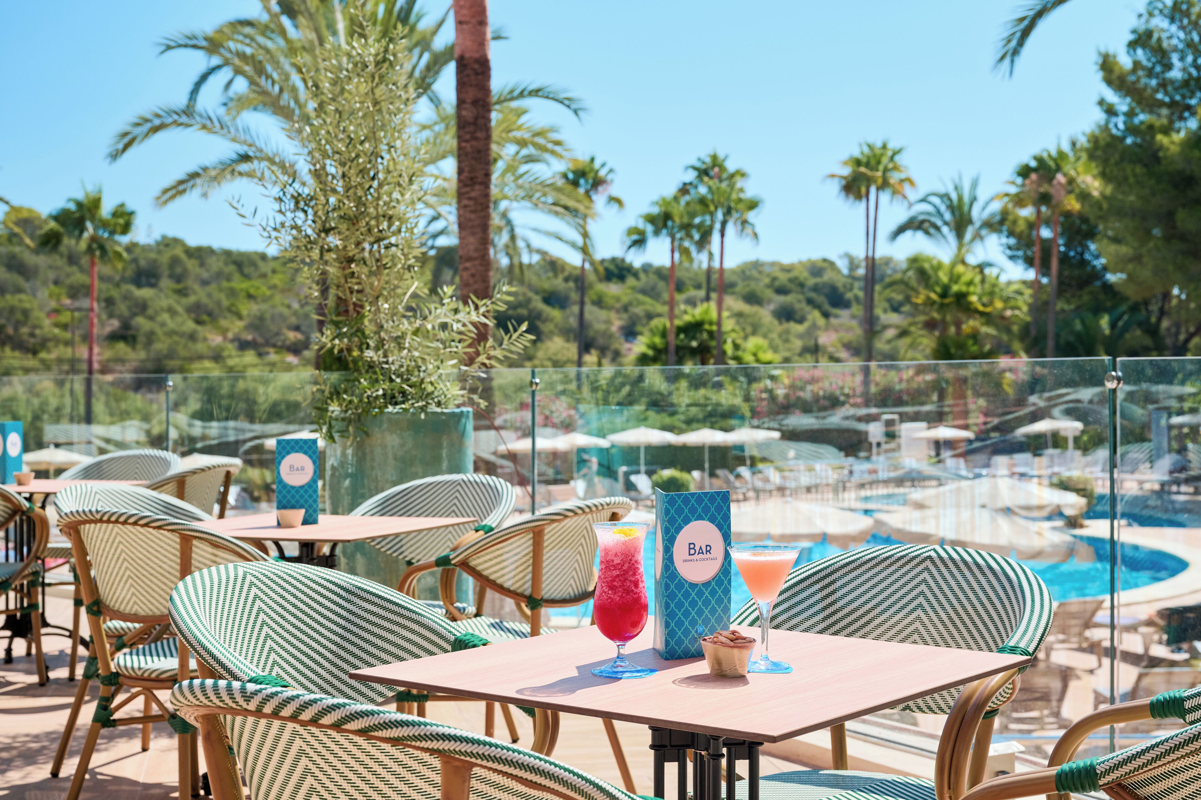 Zona de bar al aire libre con sillas y mesas de mimbre, una piscina al fondo y un expositor de menú.