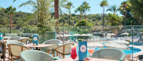 An outdoor bar area with wicker chairs and tables, a pool in the background, and a menu stand.