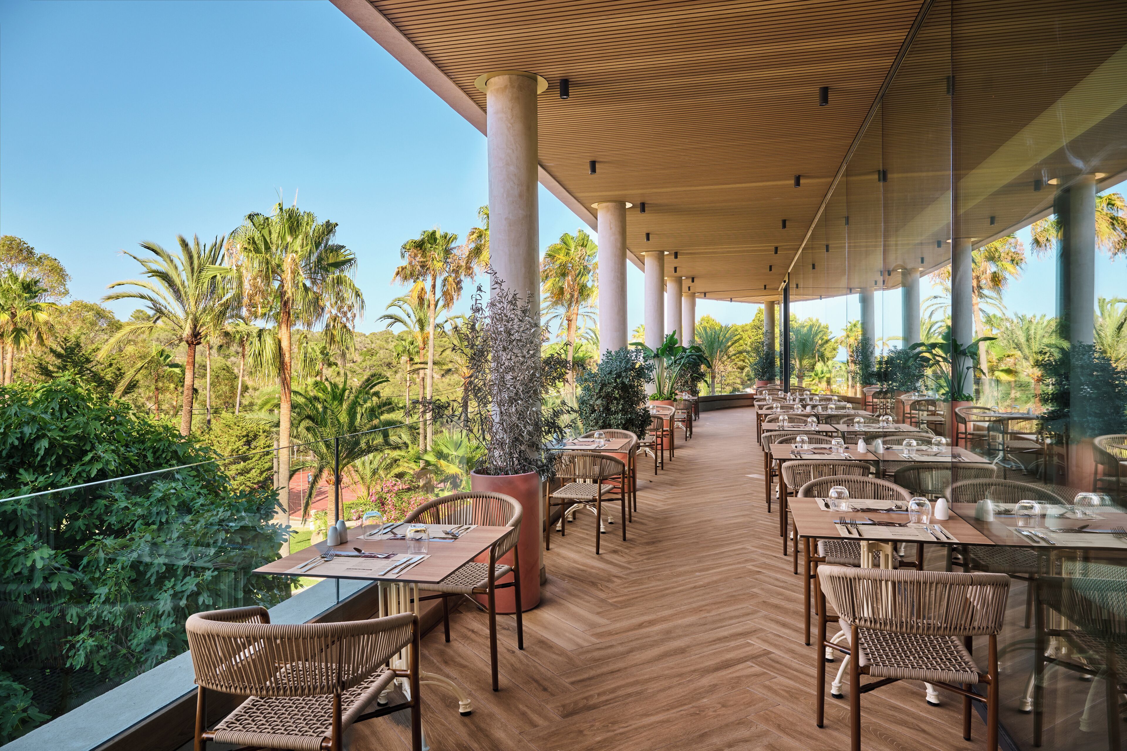 An outdoor dining area with wooden tables and chairs, surrounded by lush greenery and palm trees.