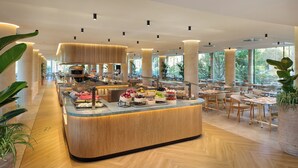 Un moderno vestíbulo de hotel con un mostrador central que exhibe diferentes productos alimenticios, rodeado de zonas de estar y grandes ventanales.