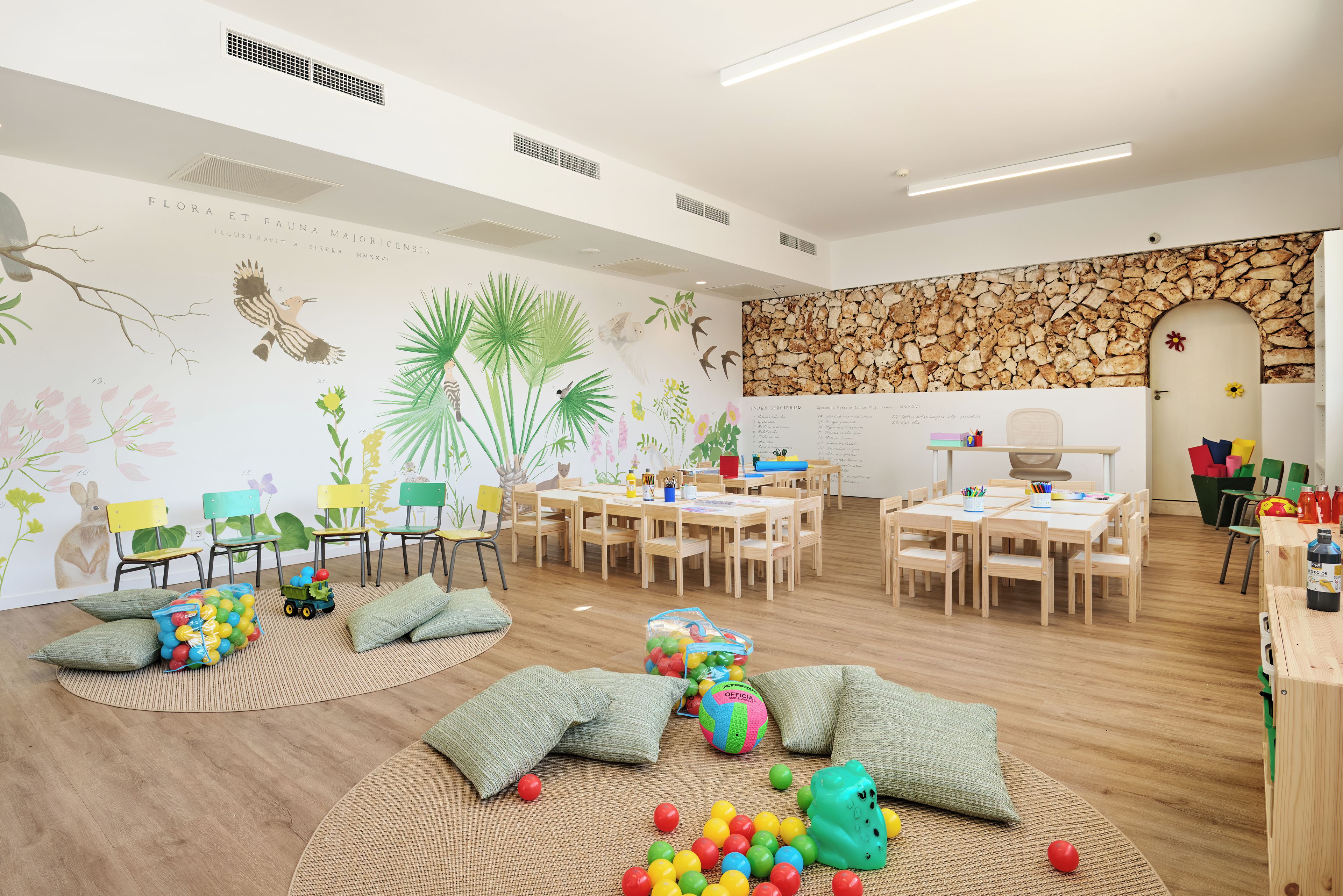A spacious room with wooden flooring, a stone wall, and a whiteboard. There are several wooden tables and chairs arranged for group activities.