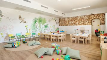 A spacious room with wooden flooring, a stone wall, and a whiteboard. There are several wooden tables and chairs arranged for group activities.