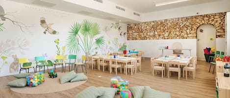 Une pièce spacieuse dotée d’un plancher en bois, d’un mur en pierre et d’un tableau blanc. On y trouve plusieurs tables et chaises en bois, disposées pour les activités de groupe.