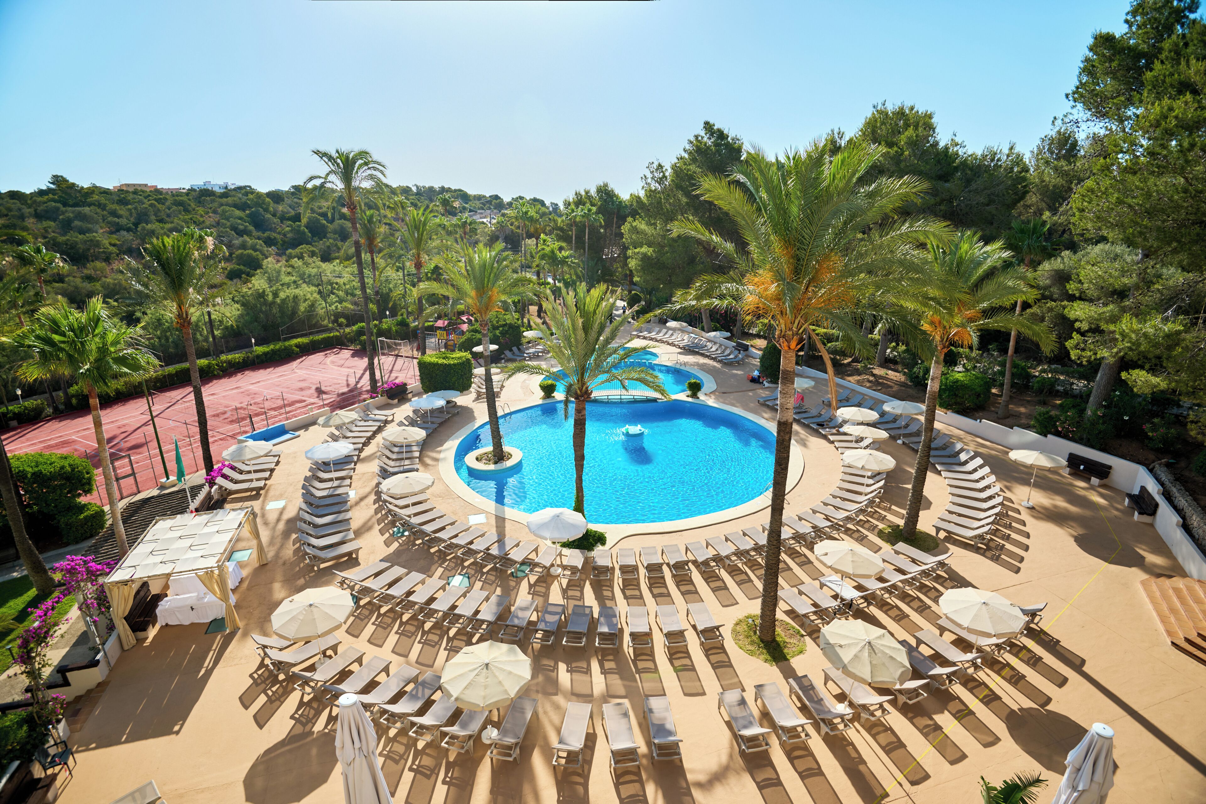 A resort with a central swimming pool surrounded by sun loungers and palm trees.