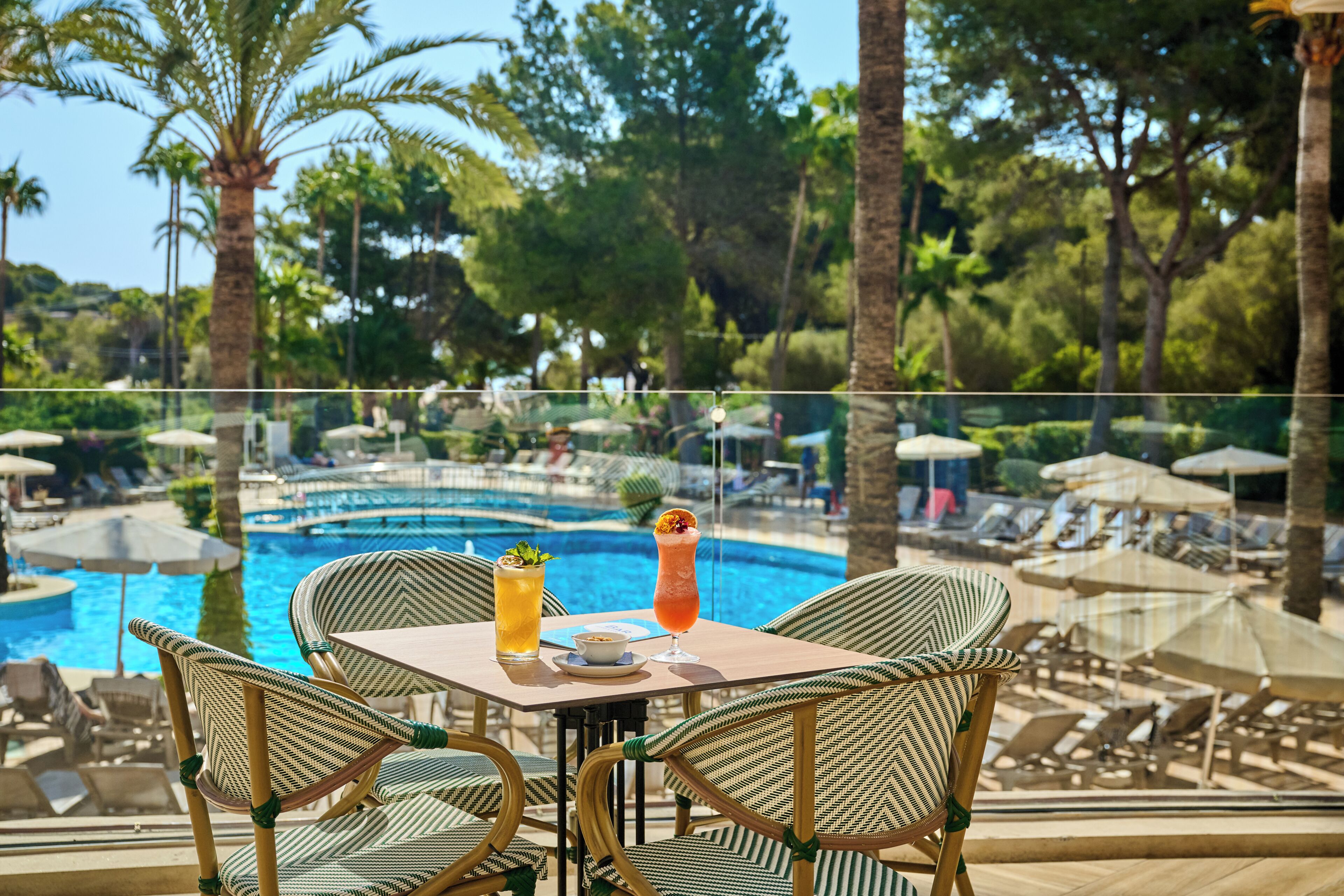 A poolside area with wicker chairs, a table with drinks, and a view of the pool and surrounding greenery.