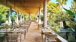 Un restaurante al aire libre con mesas y sillas de vidrio, rodeado de palmeras y follaje.