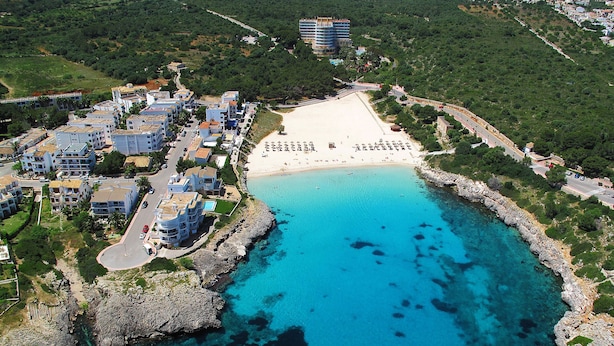 Vista aérea de una zona costera con playa arenosa, aguas azules cristalinas y una área residencial con edificios.
