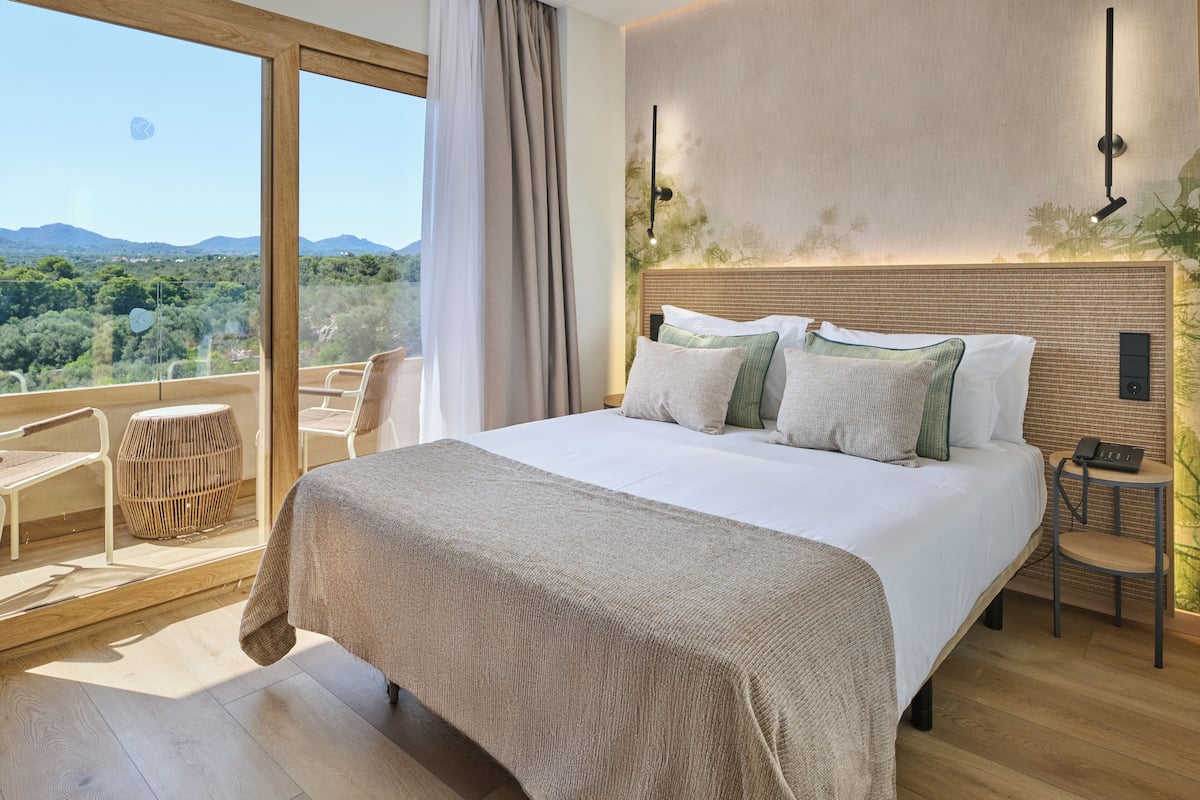 Een slaapkamer met een groot bed, uitzicht op de natuur, een klein tafeltje en een stoel.
