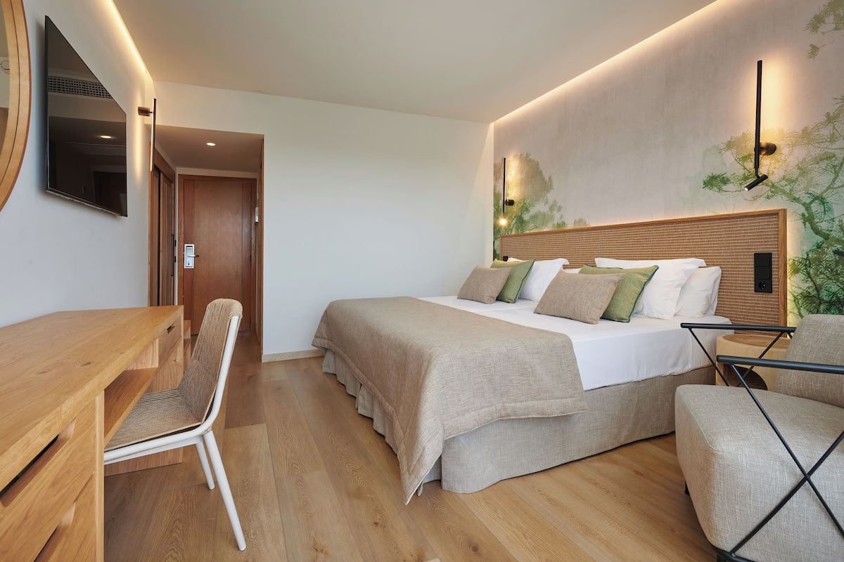 Een moderne hotelkamer met een houten bureau, een groot bed met houten hoofdbord, een stoel en een tv aan de muur.