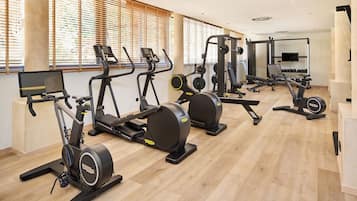 Un gimnasio con diversos equipos de ejercicio, ventanas amplias con persianas y suelo de madera.