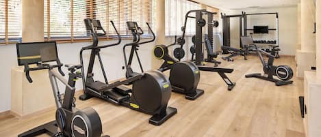 Une salle de sport équipée de divers appareils d’exercice, de grandes fenêtres avec stores et d’un plancher en bois.