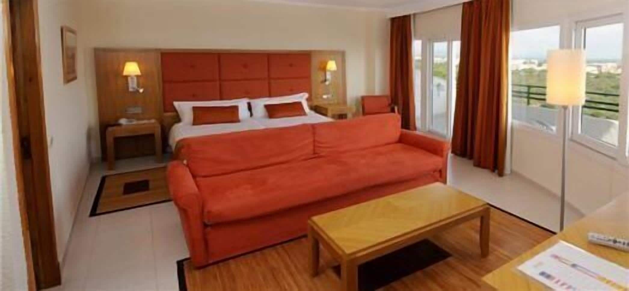 A hotel room with a large red sofa, a wooden coffee table, a bed with a red headboard, two bedside tables with lamps, and a view through the window.
