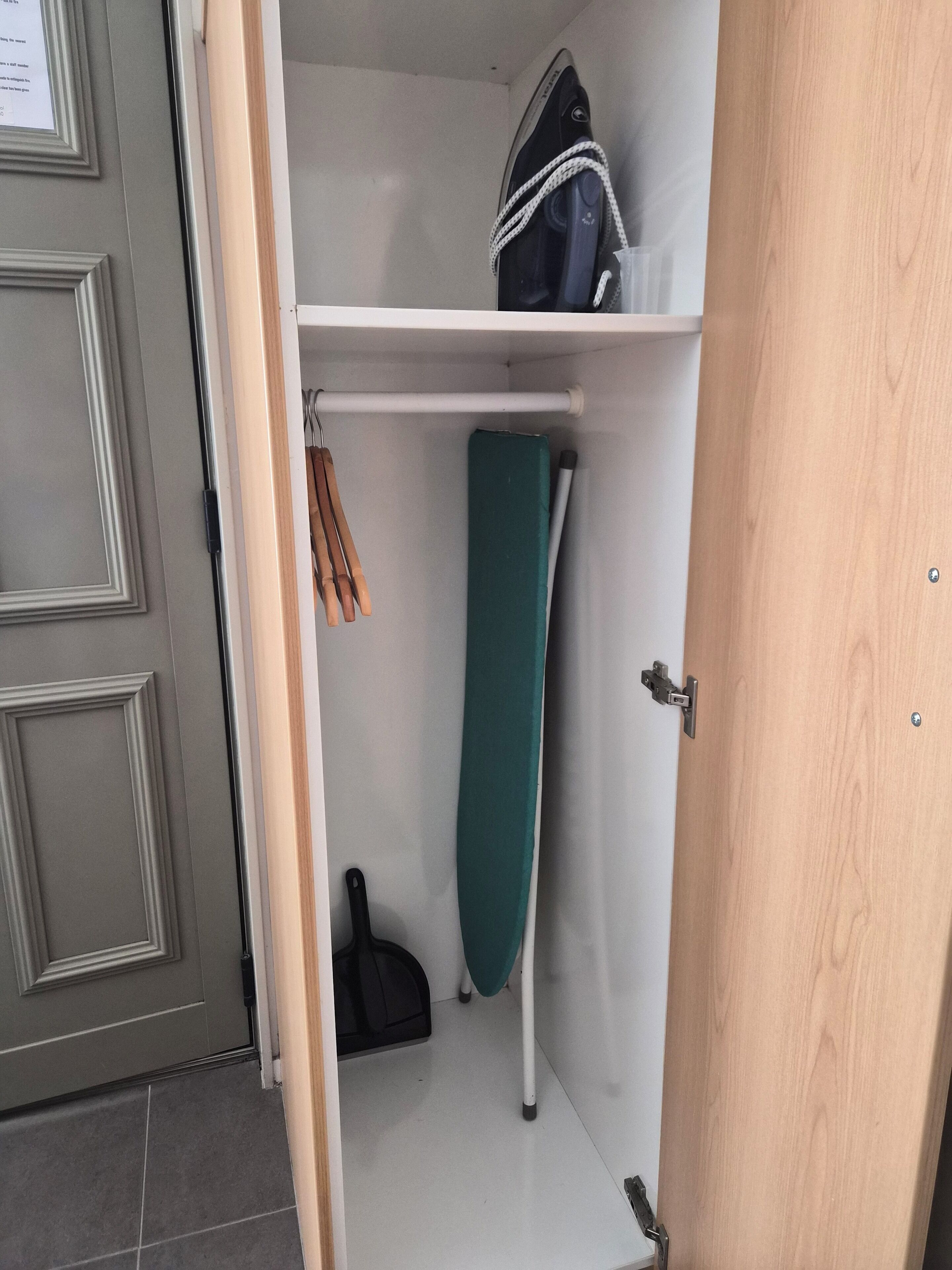 Un espacio de almacenamiento pequeño con estante, tabla de planchar y perchero.