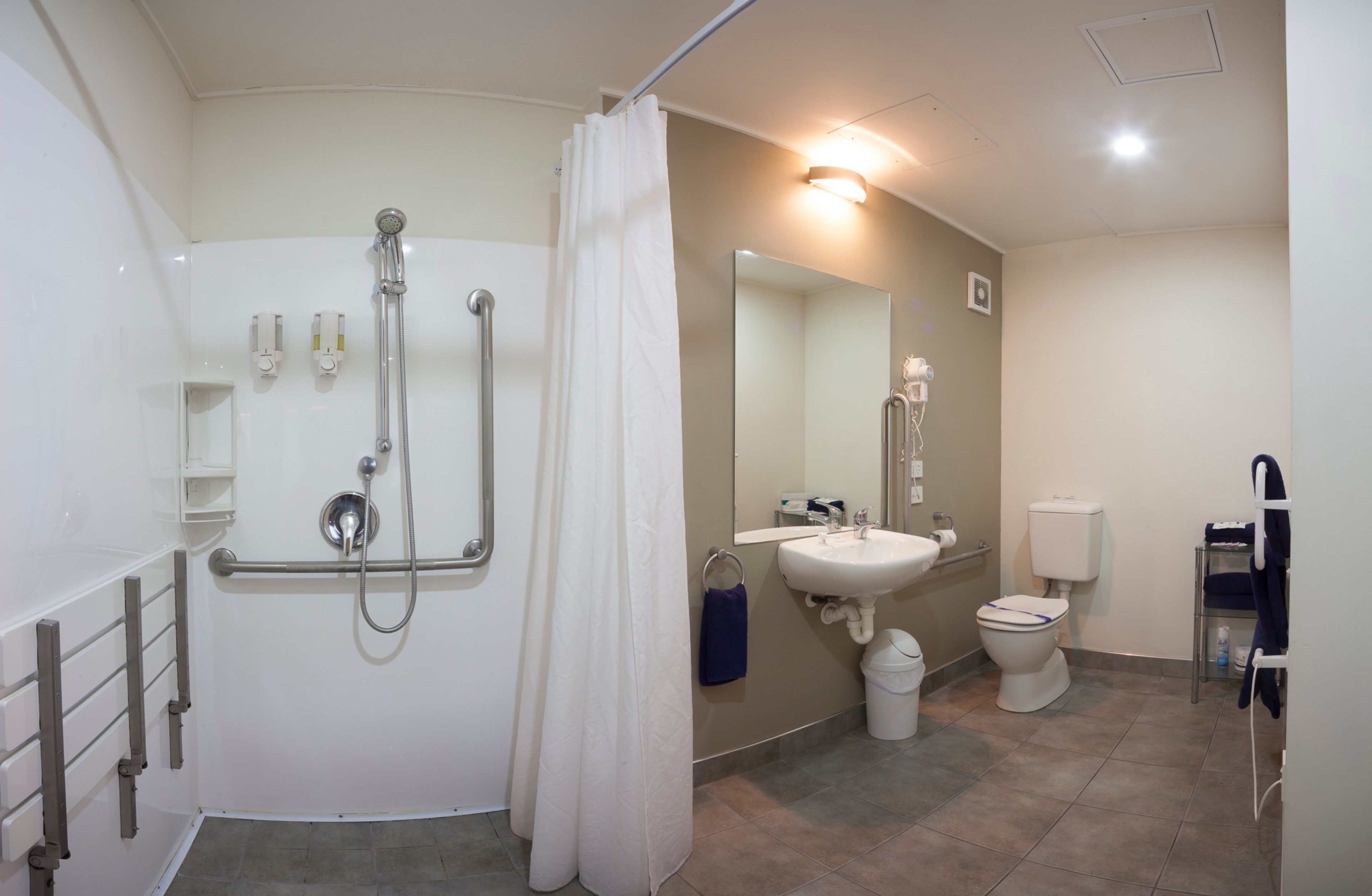 A bathroom with a shower, toilet, sink, and handrails.
