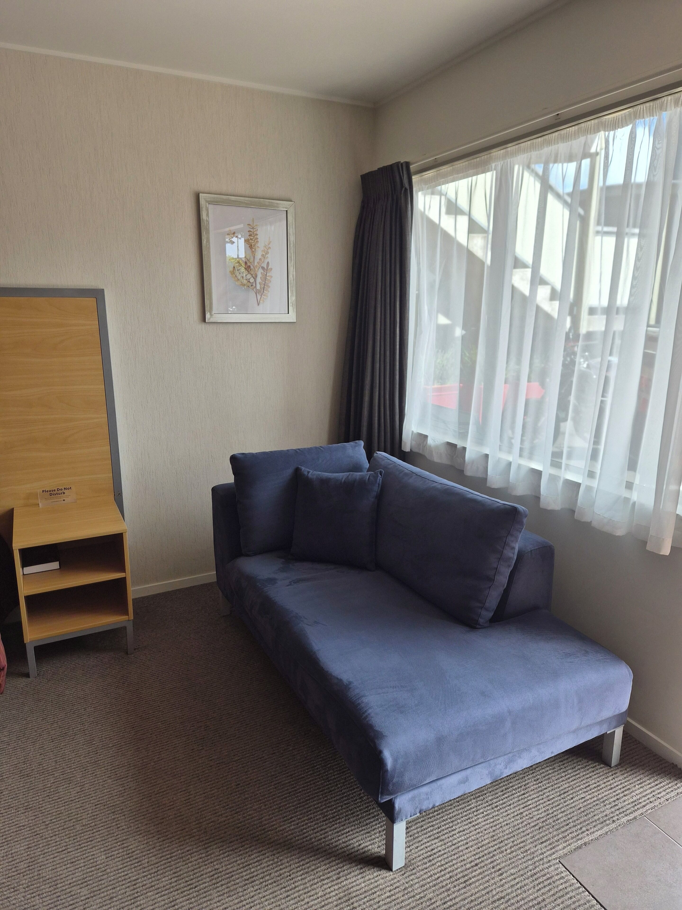 A room with a blue sofa, a small wooden cabinet, a framed picture on the wall, and a window with sheer curtains.