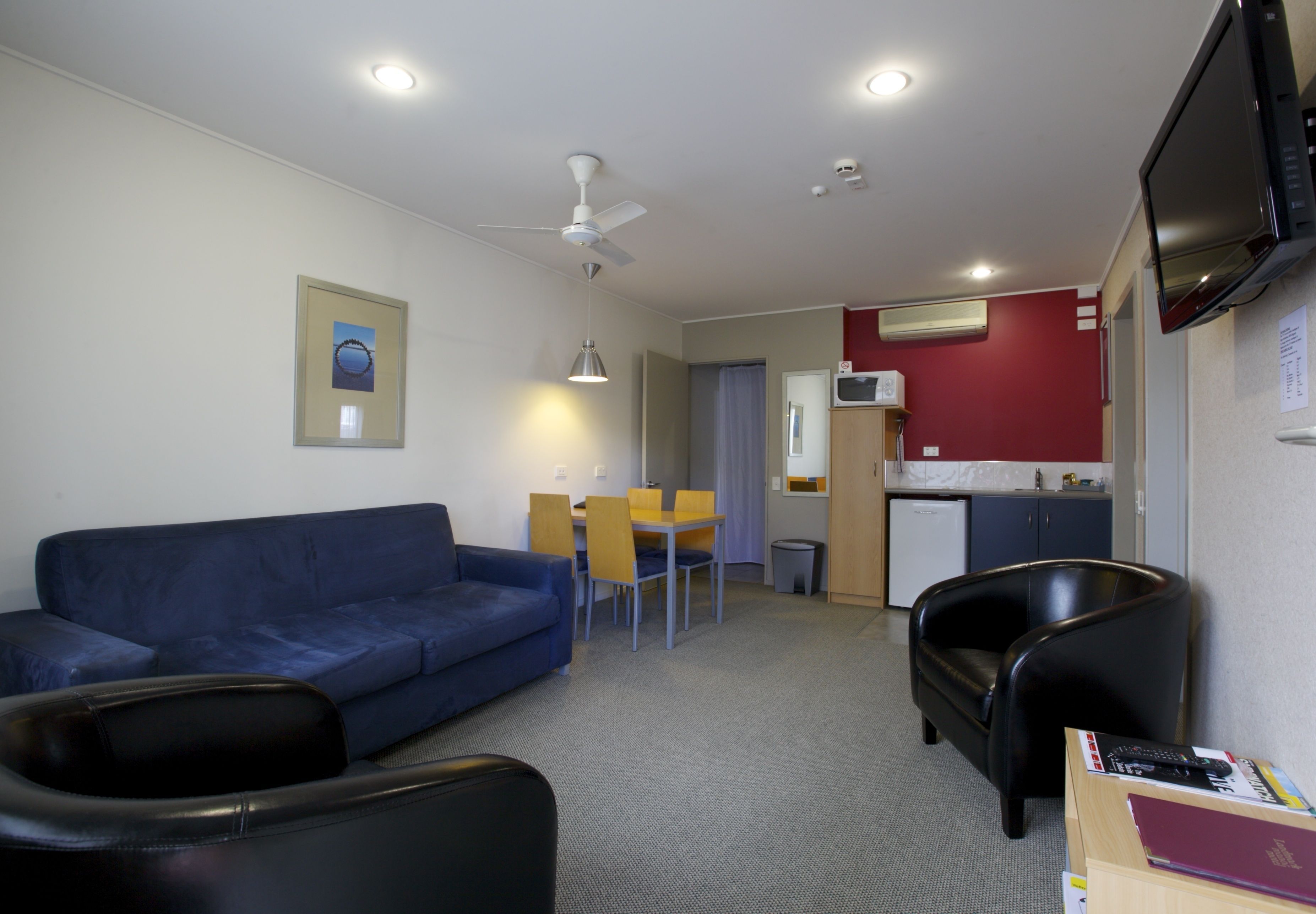 A compact living space with a blue sofa, a black armchair, a dining table with yellow chairs, a ceiling fan, recessed lighting, a TV mounted on the wall, and a small kitchenette area.