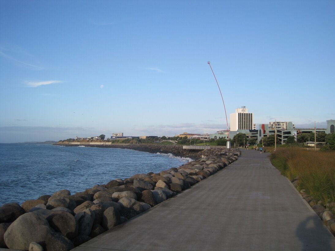 A coastal walkway with a concrete path, a rocky breakwater, and buildings in the background.