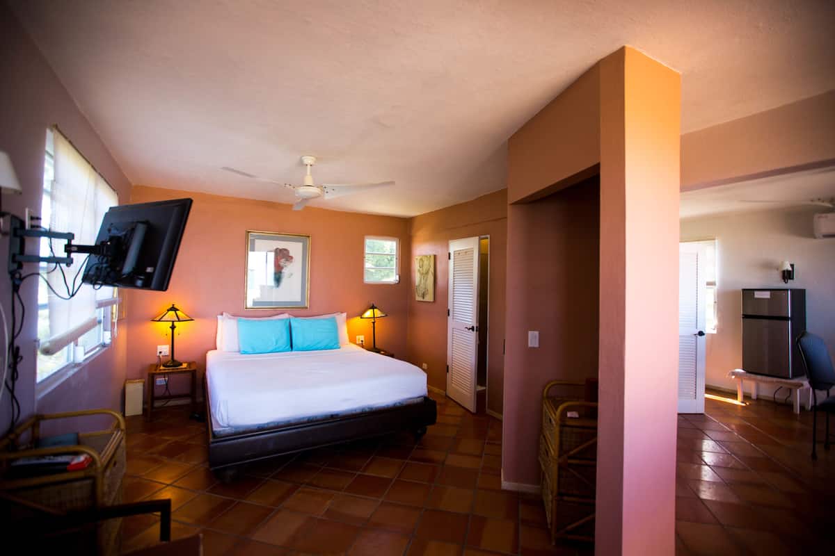 Ein Schlafzimmer mit einem Bett, zwei Nachttischlampen, einem Deckenventilator, einem an der Wand montierten Fernseher und einem Fenster mit Jalousien.