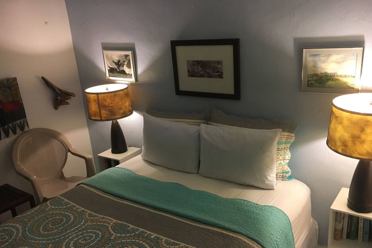 Ein ordentlich bezogenes Bett mit einem gemusterten Überwurf, zwei Nachttischlampen, ein Sessel und gerahmte Bilder an der Wand.
