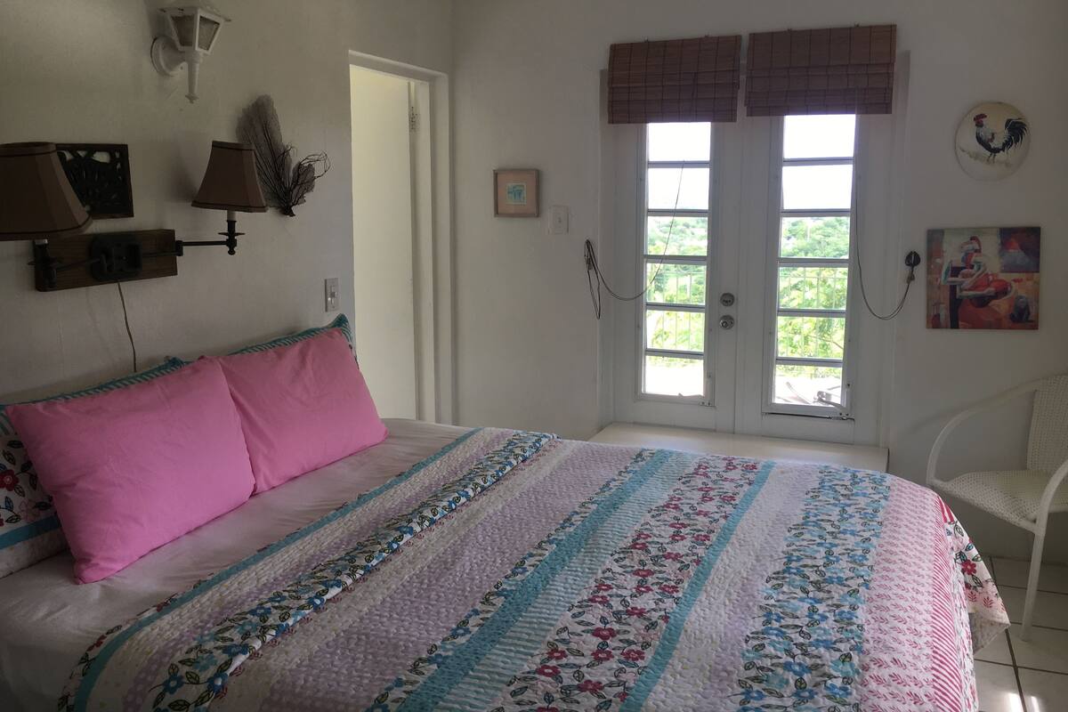 Ein Schlafzimmer mit Bett, Nachttisch, Lampe, einem Fenster mit Jalousien und einer Tür.