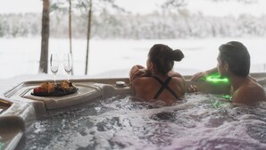Ein Whirlpool mit zwei Personen, einer Serviertablett mit Speisen und Getränken und Blick auf eine verschneite Landschaft.