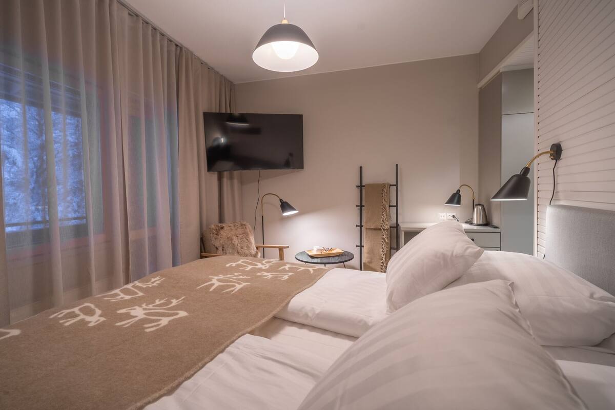 Ein modernes Schlafzimmer mit Bett, einem an der Wand montierten Fernseher, einem Schreibtisch mit Lampe und einem Fenster mit Vorhängen.