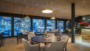Ein moderner Essbereich mit runden Tischen und Stühlen, großen Fenstern mit Blick auf Winterlandschaften und einer Bar mit Weinkühlschrank.