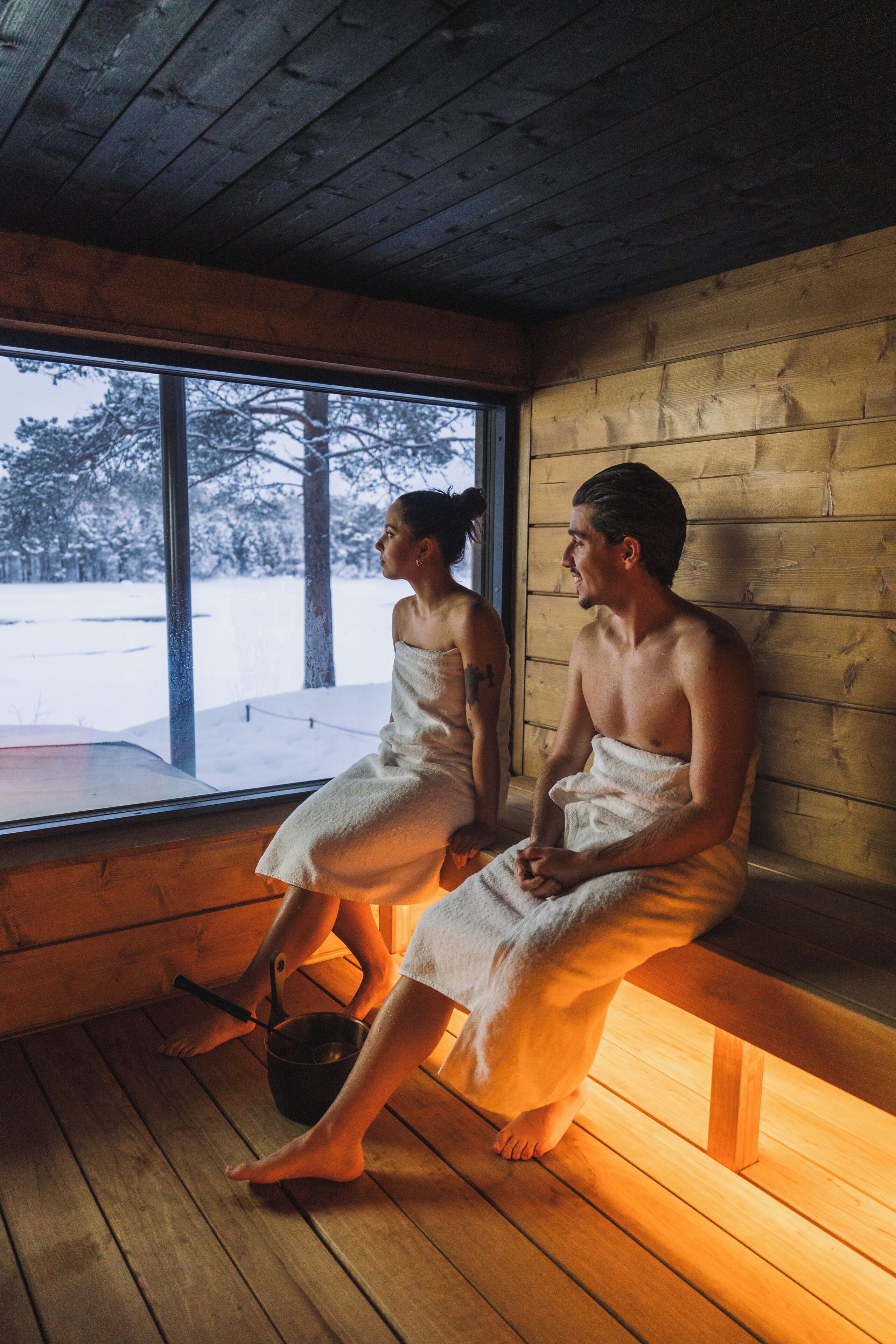 Two people sitting on a bench inside a wooden cabin with a window.