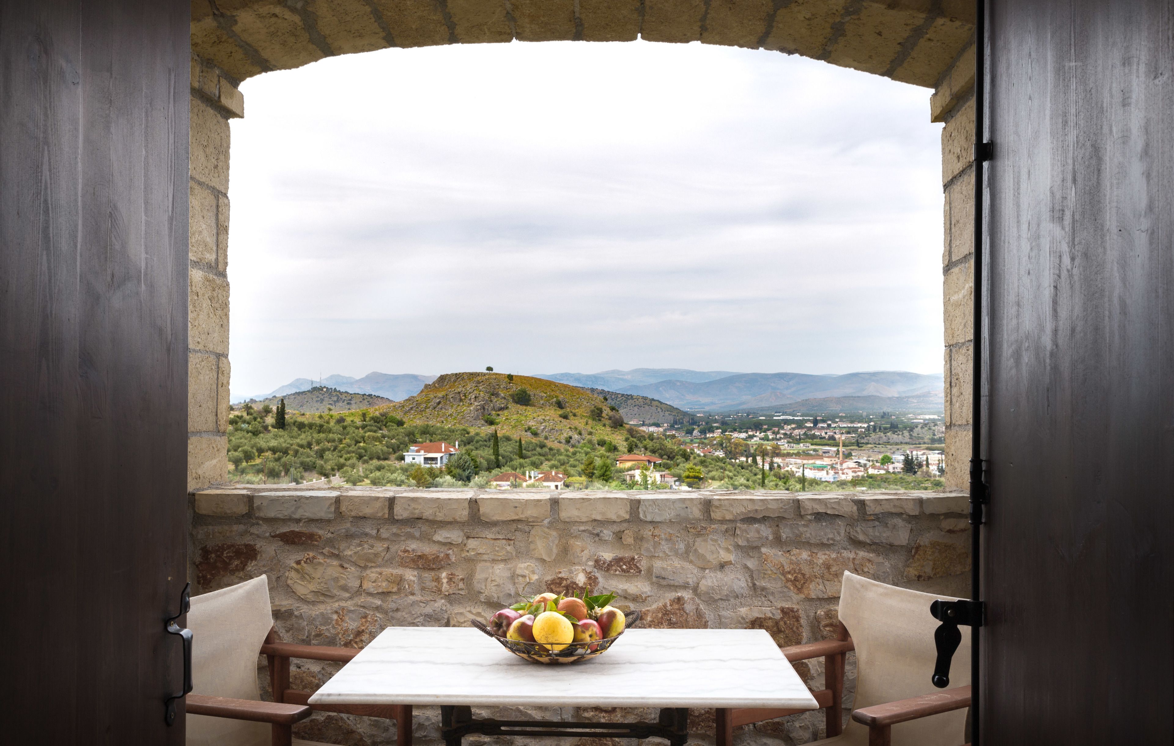 A table with a fruit bowl, two chairs, and a stone wall with a view of a hilly landscape.