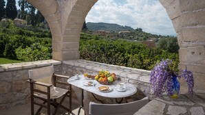 Una mesa de piedra con un frutero, platos y tazas, frente a un arco de piedra con vistas a un paisaje exuberante.