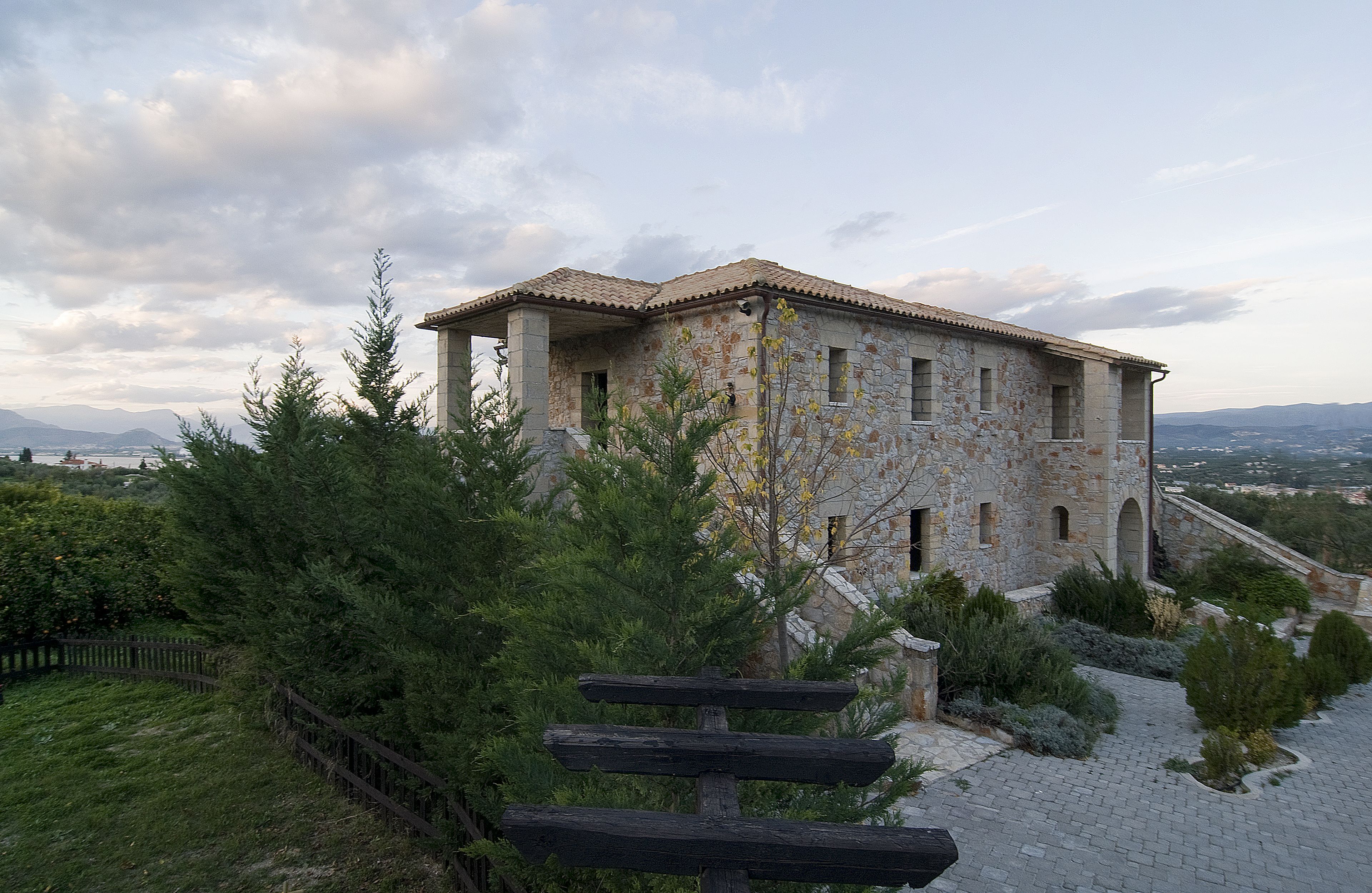 Un bâtiment en pierre, doté d’un toit en tuiles, entouré de verdure et bordé d’un chemin pavé.