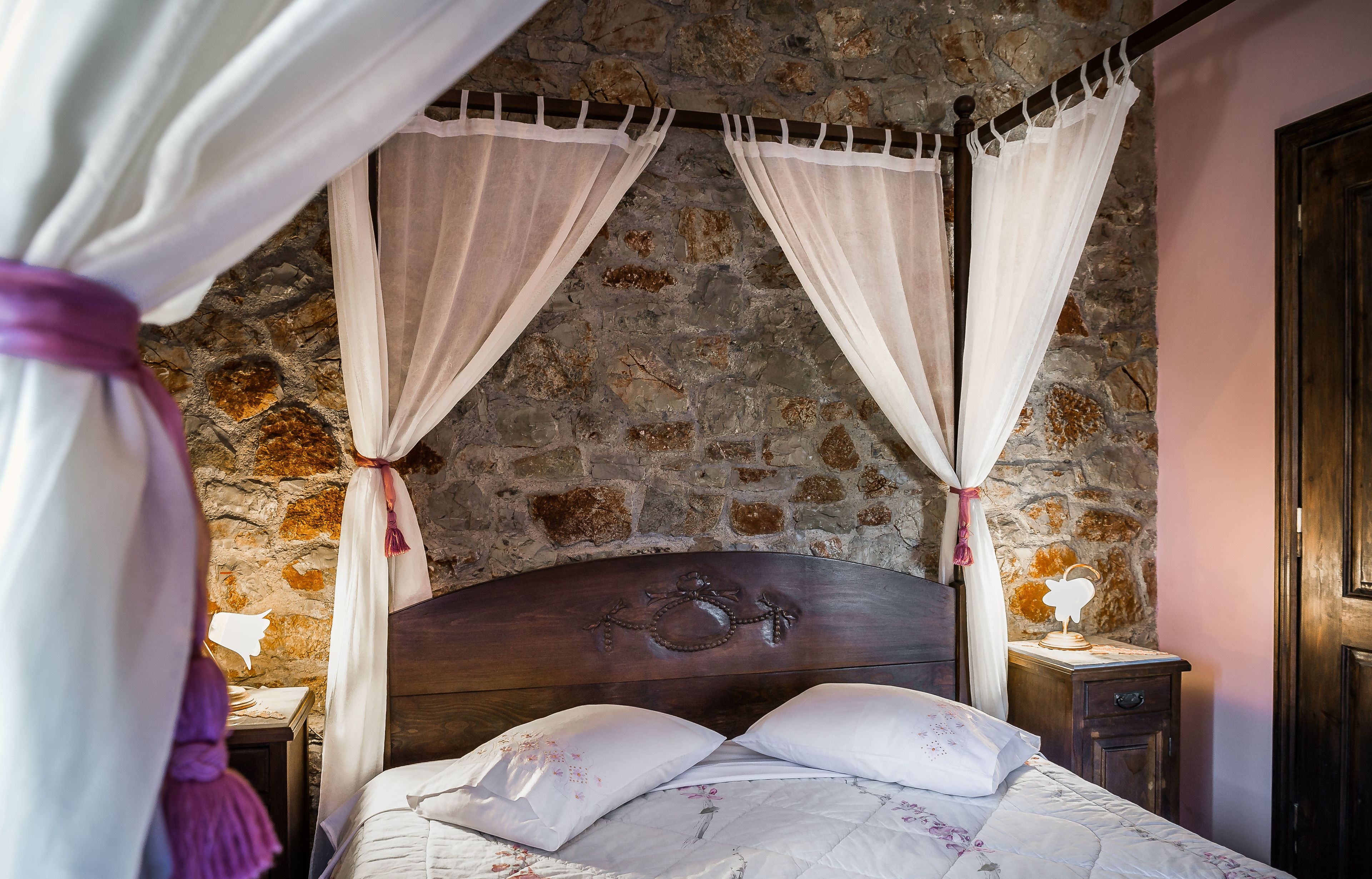 Un dormitorio con pared de piedra, cama de madera con cabecero tallado y mesita de noche con lámpara.