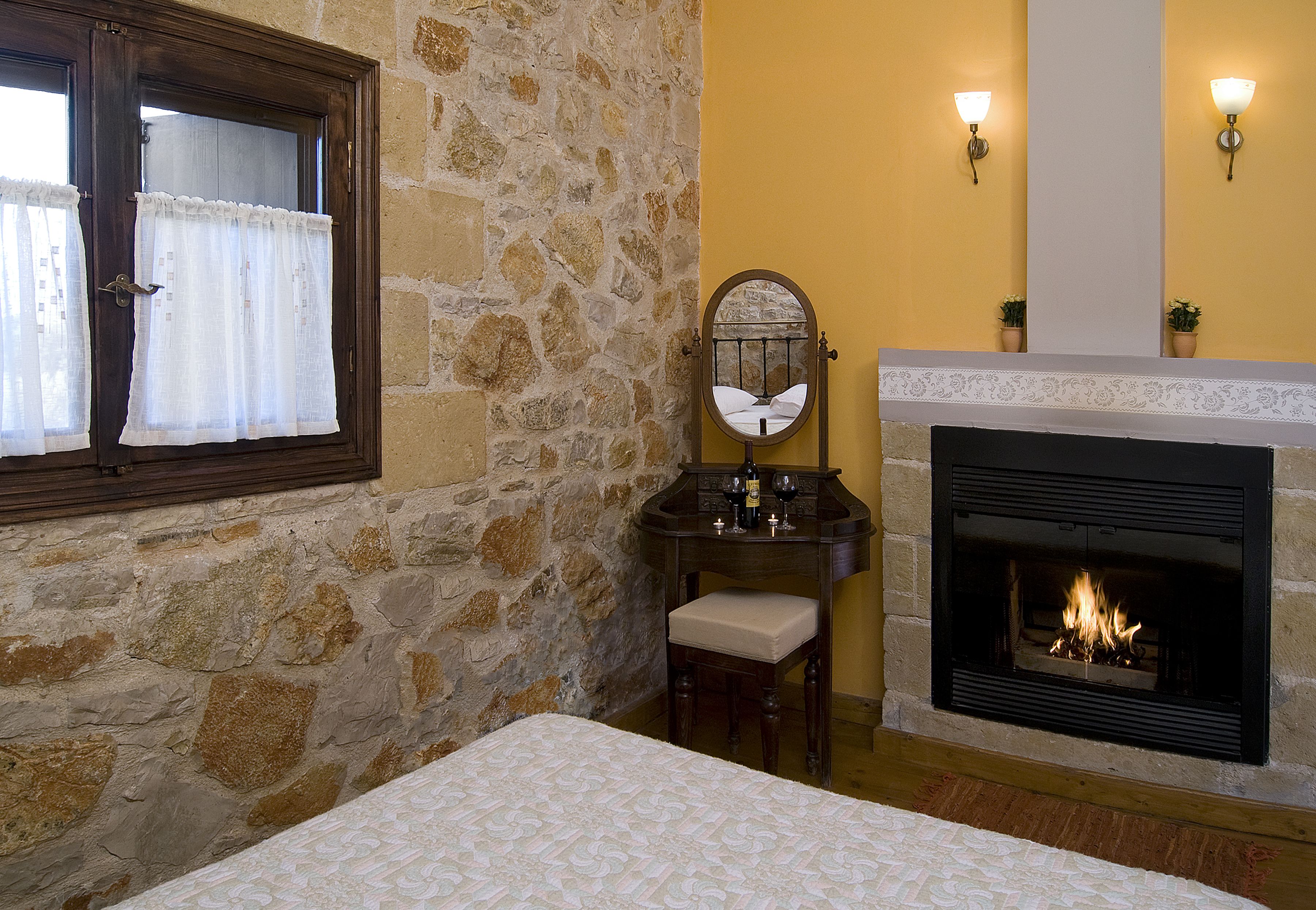 A bedroom with a stone wall, a fireplace, a bed, a nightstand, a mirror, and a window with curtains.