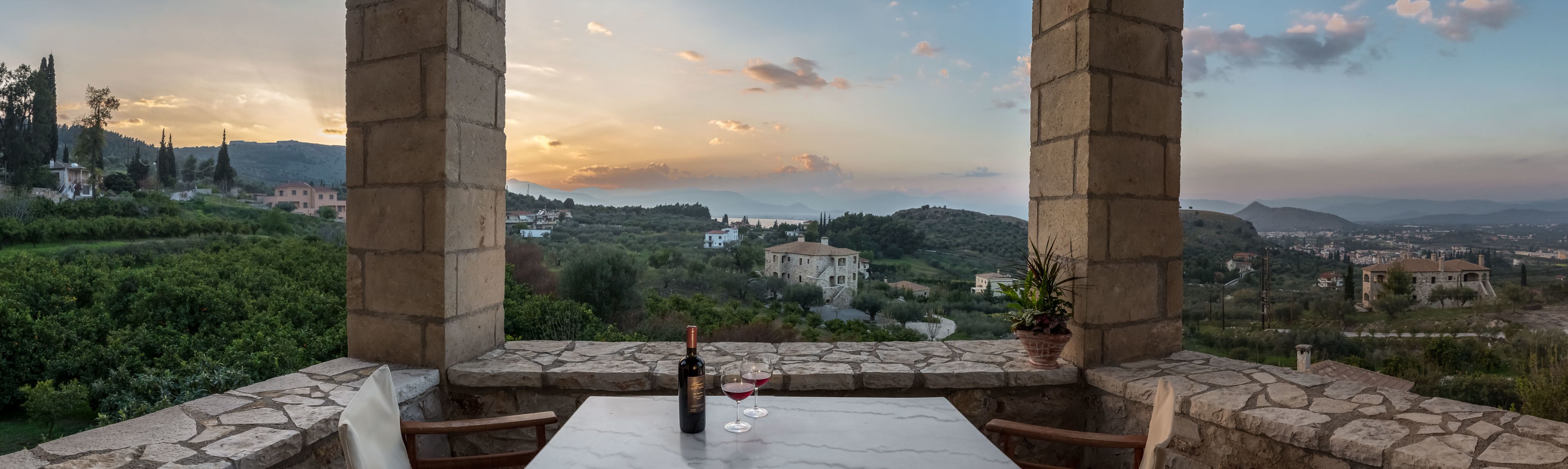 A terrace with a table, wine bottle, and glasses, overlooking a scenic landscape with buildings and hills.