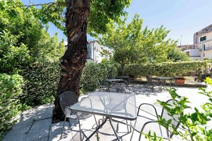 An outdoor patio with metal tables and chairs, surrounded by greenery and a tree.