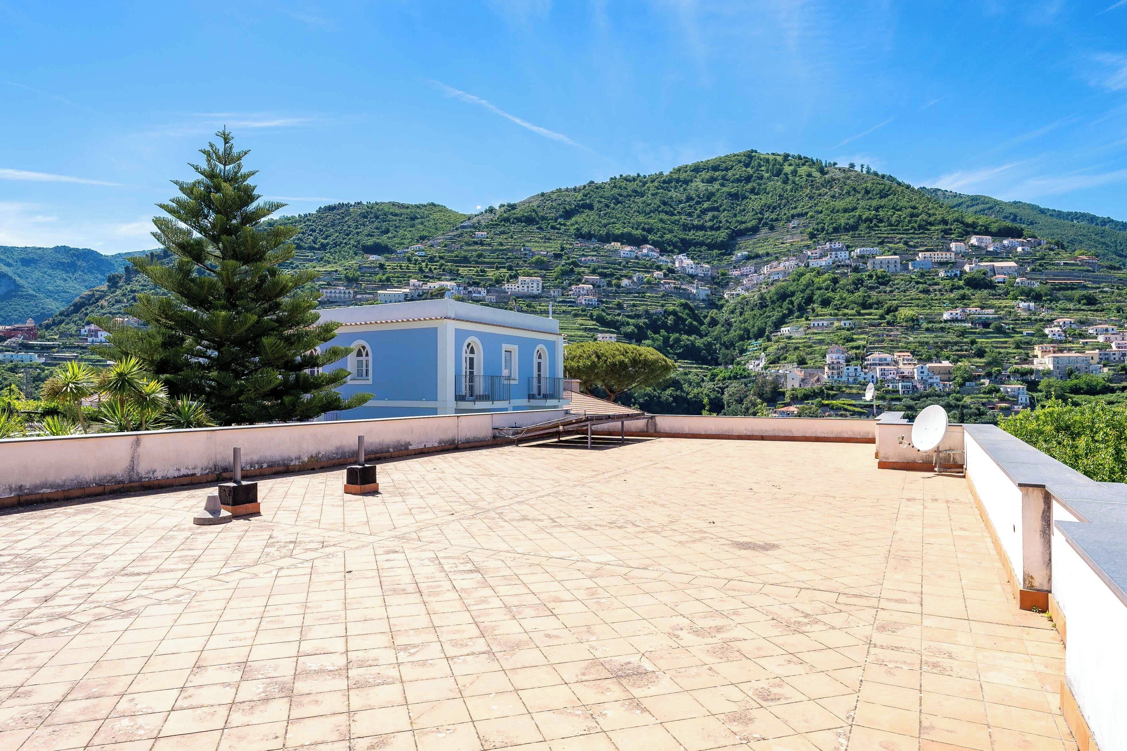 A rooftop terrace with a view of a hillside village, a blue building, and a satellite dish.