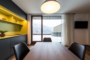 Uma área de jantar moderna com mesa e cadeiras de madeira, uma estante embutida com plantas, uma grande janela com vista para montanhas e uma televisão de tela plana fixada na parede.