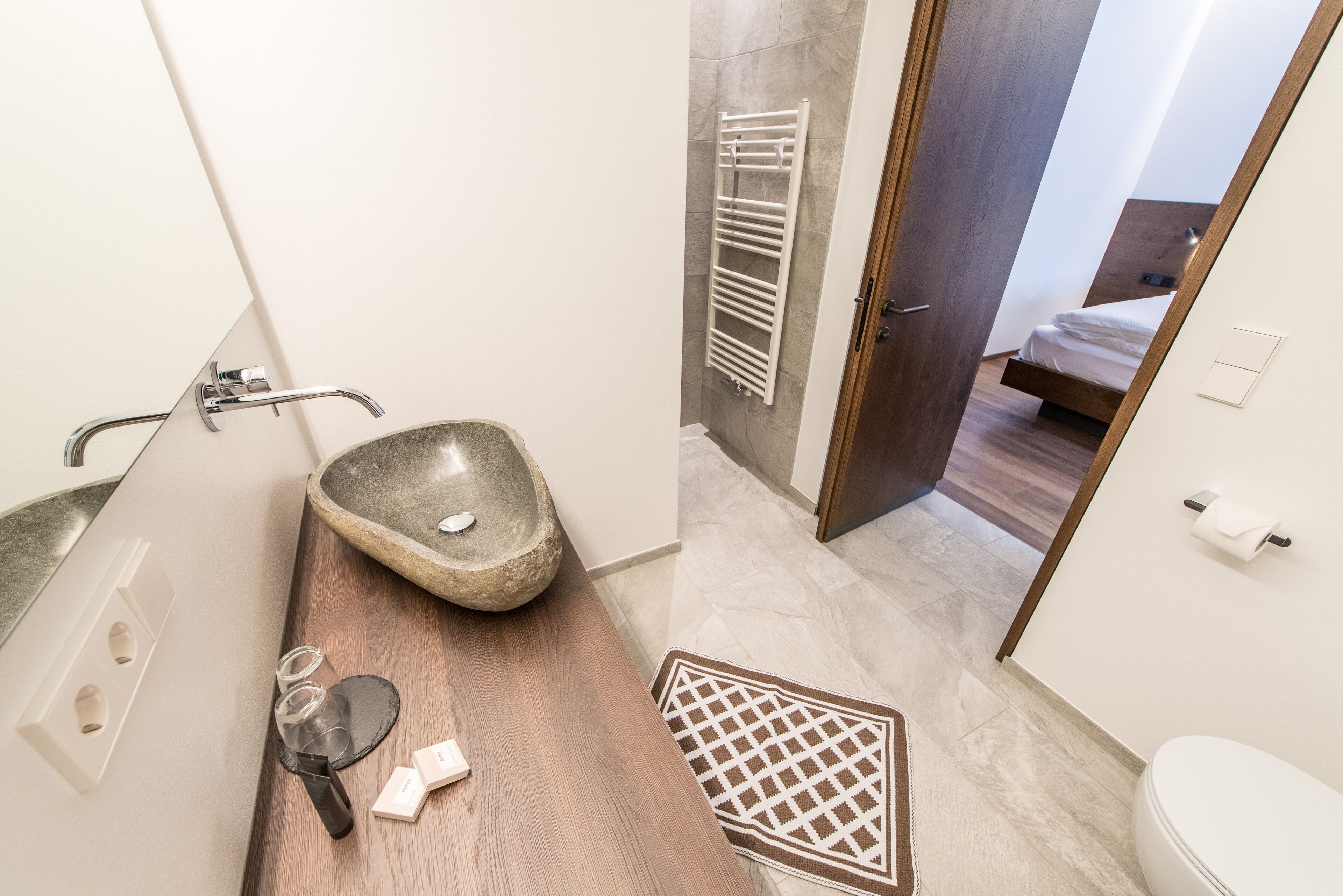 A modern bathroom with a stone sink, a wooden floor, a white toilet, and a visible bed in the adjacent room.
